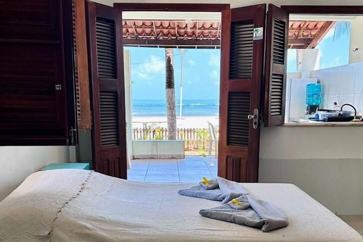 Quarto de hotel com cama, vista para a praia e área de cozinha compacta.
