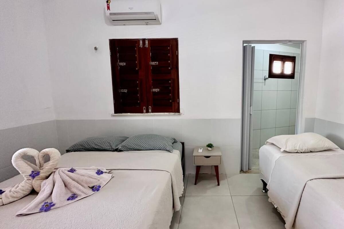 Quarto de hotel com duas camas, uma mesa de cabeceira pequena, ar condicionado fixado na parede e uma porta que dá para outro quarto.