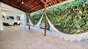 Un espacio al aire libre cubierto con una hamaca, suelo de baldosas, sillas y una pared de vegetación.