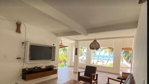 Un salón amplio con televisor de pantalla plana en la pared, muebles de madera y ventanas grandes con vistas a la playa.