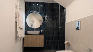 Un baño moderno con un lavamanos blanco, paredes cubiertas de baldosas oscuras, un espejo redondo y un inodoro.