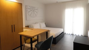 Une chambre d’hôtel moderne avec une armoire en bois, un lit avec du linge de maison blanc, un bureau avec une chaise et une grande fenêtre avec des rideaux légers.