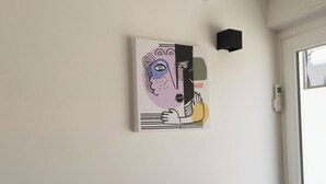 Een kamer met een houten tafel, een zwarte stoel, een wandlamp en een abstract kunstwerk.