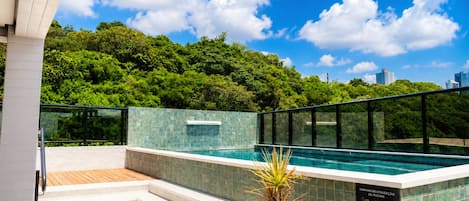 A rooftop swimming pool with a glass balustrade, surrounded by lush greenery and a clear blue sky.