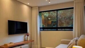 A modern living room with a flat-screen TV, a sofa, a small table, and a window with curtains. - Mahalo Flat - LikeHome Hospedagens (Joao Pessoa)