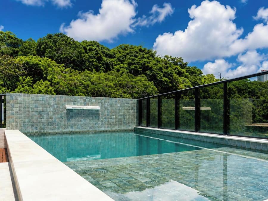 Una piscina con paredes de vidrio, rodeada por una terraza con baldosas y vistas a vegetación exuberante.