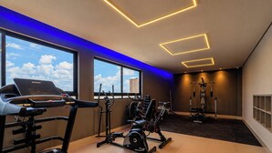 A modern gym with cardio equipment, weight machines, and a large window offering a city view. - Mahalo Flat - LikeHome Hospedagens (Joao Pessoa)