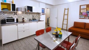 Pieni, käytännöllinen keittiö, jossa on valkoiset kaapit, mikroaaltouuni ja pesuallas. Keittiössä on ruokapöytä tuoleineen ja maljakko, jossa on kukkia. Punainen tuoli on näkyvissä, ja seinää vasten on nojaamassa tikkaat.