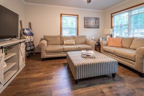 A living room with a beige sofa, a striped ottoman, a television, and a window with blinds.