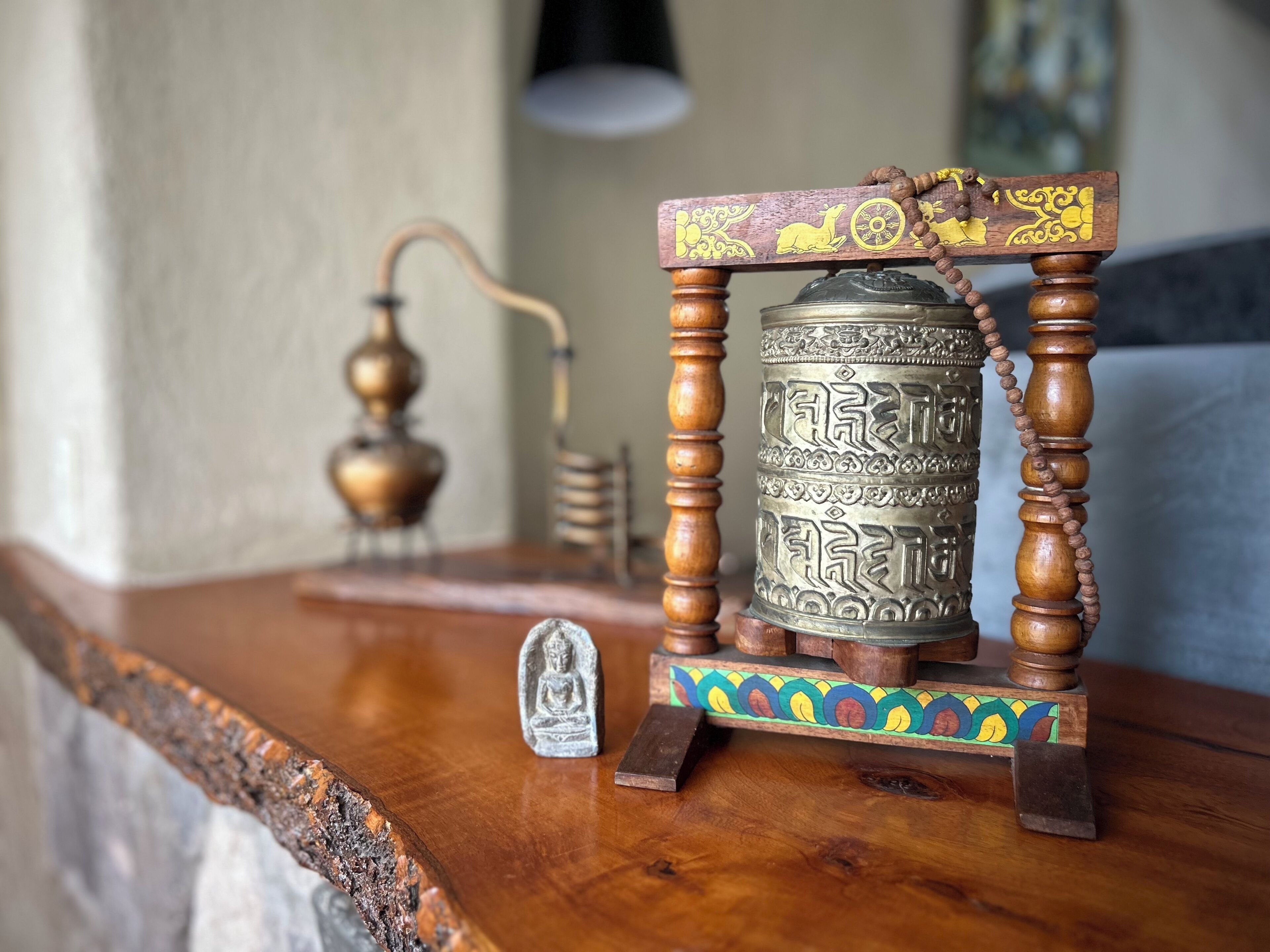 Una mesa de madera con un objeto metálico encima, una pequeña estatua y una lámpara al fondo.