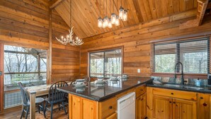 A wooden cabin interior with a kitchen area featuring a wooden island, a chandelier, and a view through large windows. - Ellijay Mountain View, Pets, Pool, Firepit, HotTub, Gameroom, Grill & Fireplace (Ellijay)