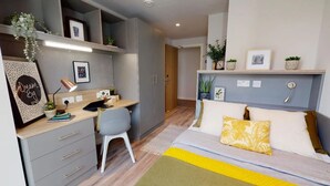 Una habitación compacta con una cama, un escritorio y una silla. Hay estanterías con plantas y cuadros enmarcados.