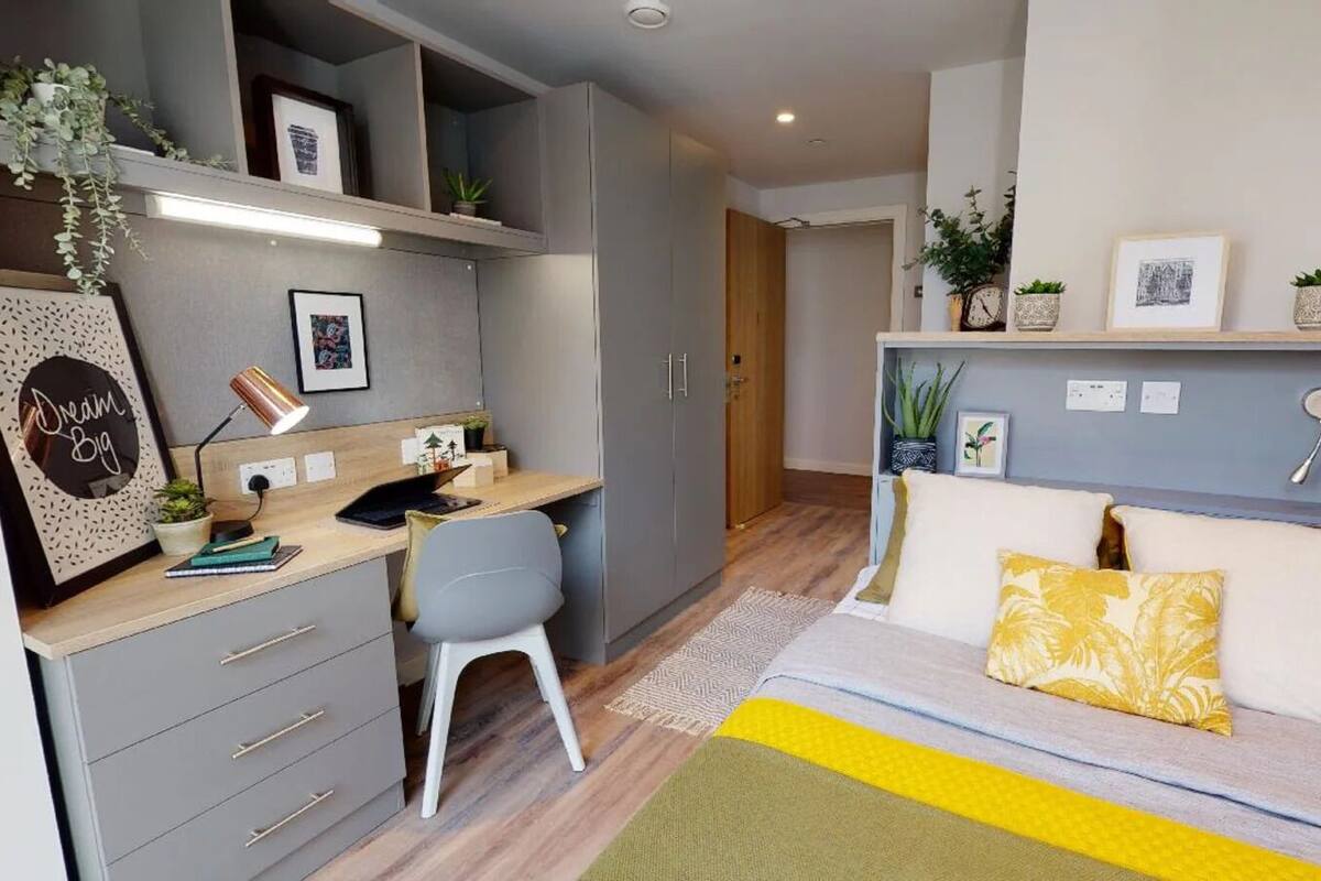 A compact bedroom with a bed, a desk, and a chair. There are shelves with plants and framed pictures.