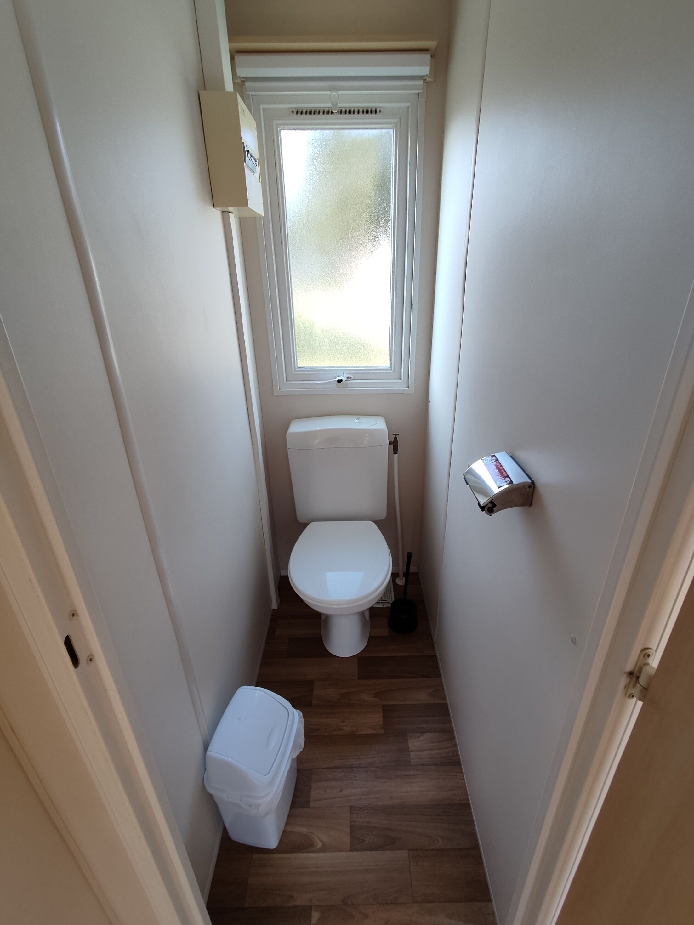 Ein kleines, schmales Badezimmer mit Toilette, Fenster, Besen und Mülleimer.