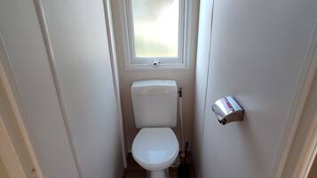 Een kleine, smalle badkamer met toilet, raam, bezem en prullenbak.