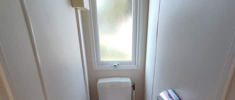 A small, narrow bathroom with a toilet, a window, a broom, and a trash bin.
