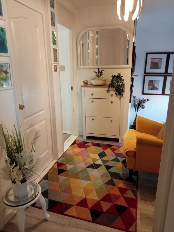 Un couloir avec une porte blanche, un meuble blanc avec des tiroirs, un tapis géométrique aux couleurs vives, une petite table avec une plante et un fauteuil jaune.