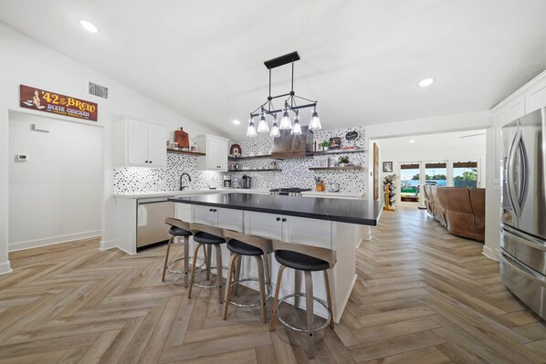 A modern kitchen with a central island, bar stools, a stainless steel refrigerator, and a sign for '42 & Brews College Station, TX'.