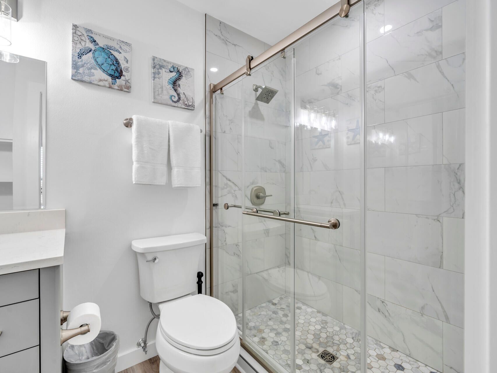 Une salle de bain moderne avec une douche à parois de verre, des carreaux de marbre blanc et deux œuvres d’art accrochées au mur.