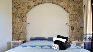 Ein Schlafzimmer mit Steinsäulenbogen, einem Bett mit gemustertem Überwurf, zwei Kissen und einem Nachttisch mit Lampe und Büchern.