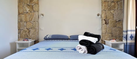 A bedroom with a stone wall arch, a bed with a patterned bedspread, two pillows, and a nightstand with a lamp and books.