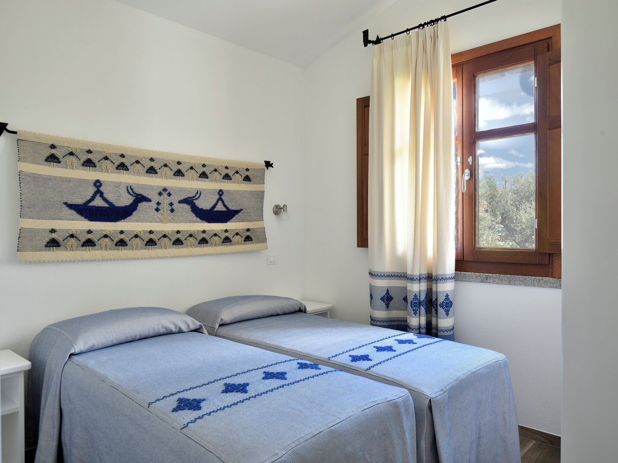 A room with two beds, a window with curtains, a wall hanging, and a view of trees outside.