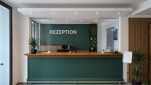 Een receptiebalie met een groene wand, een bord met de tekst 'RECEPTION', een computer en een potplant.