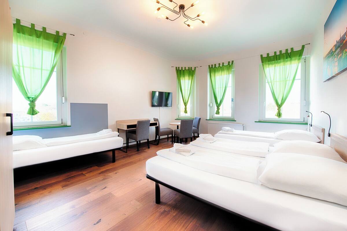 Une chambre d’hôtel avec trois lits, un bureau, des chaises, une télévision et des rideaux verts.