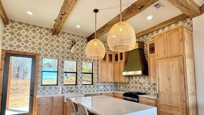 A spacious kitchen with wooden beams, a large island, pendant lights, and a view of the outdoors through the windows. - Sunday House at The Sanctuary (Gillespie County)