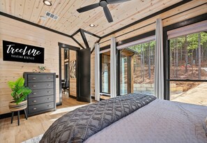 A bedroom with a large bed, a desk, and a chair, with a view of a forest through large windows.