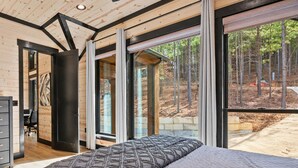 Un dormitorio con una cama grande, un escritorio y una silla, y vistas a un bosque a través de ventanas amplias.