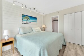 A bedroom with a bed, a nightstand, a lamp, a painting of a beach scene, and a wardrobe. - Steps to the Sand, Endless Ocean Views – Book Now! 2 bedroom, 2 full bath! (Myrtle Beach)