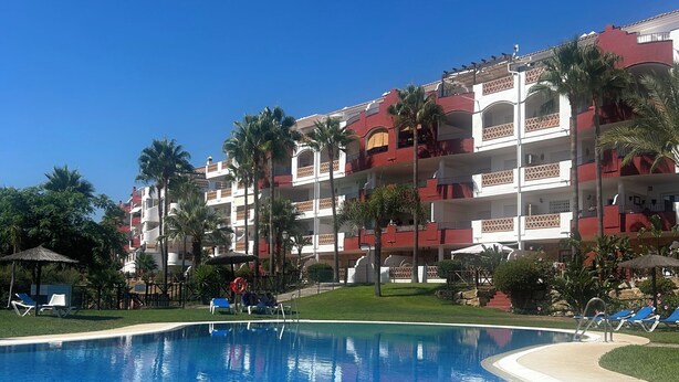A swimming pool with clear blue water, surrounded by lounge chairs and palm trees, with a building featuring balconies in the background.