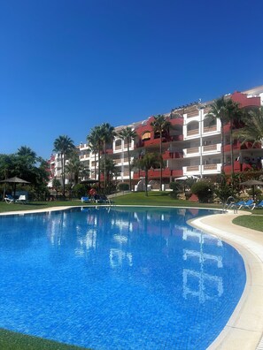 Una piscina con agua azul cristalina, rodeada de sillas de descanso y palmeras, con un edificio de balcones al fondo.
