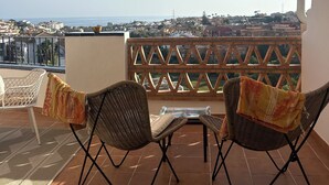 A terrace with two wicker chairs, a small table, and a view of the city and sea.