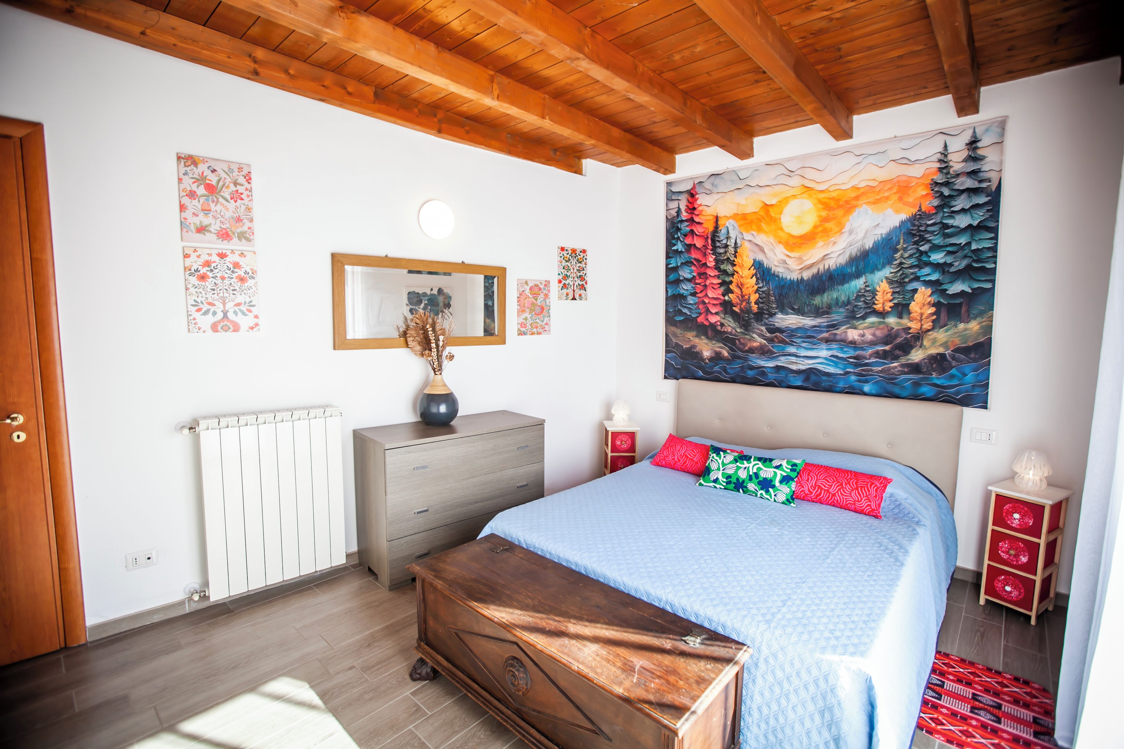 Une chambre avec un plafond en bois, un lit avec une literie bleue, un tableau représentant une scène de forêt et un radiateur.