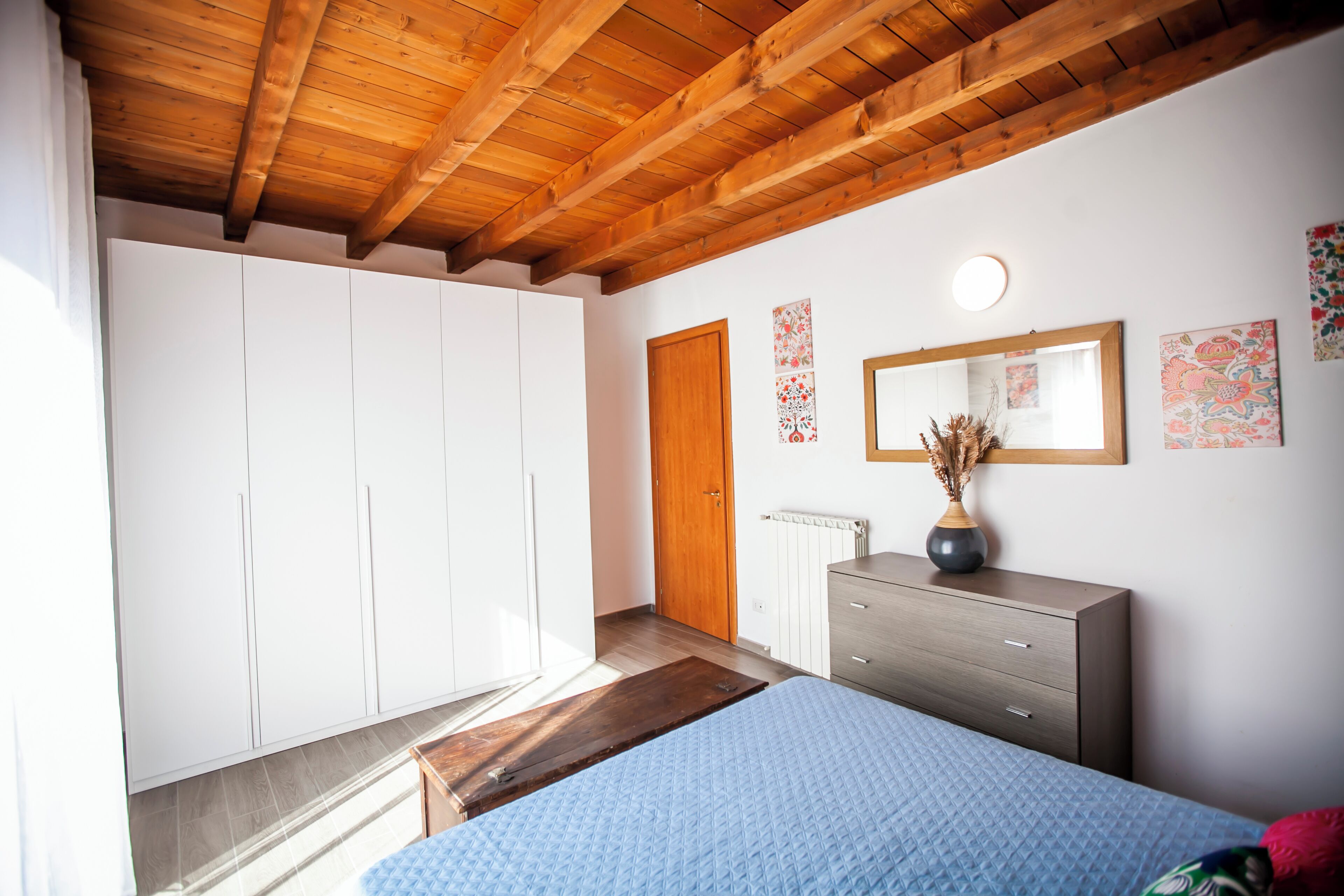 A bedroom with a wooden ceiling, a bed with a blue quilt, a dresser, a mirror, and a radiator.