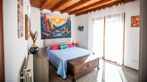 A bedroom with a wooden ceiling, a bed with a blue bedspread, a painting above the bed, and a window with curtains. - Il Borghetto (Alta Valle Intelvi)