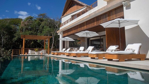 Un área junto a la piscina con sillones de descanso y sombrillas, una pérgola y un edificio con balcón de madera.