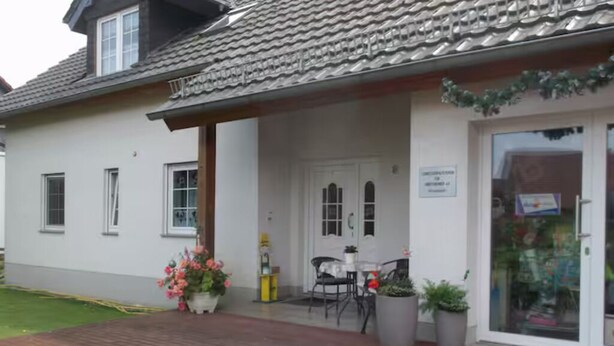 Un bâtiment avec un toit recouvert de tuiles, une façade blanche et un chemin en briques menant à une porte. On y trouve des plantes en pot et une table avec des chaises à l’extérieur.