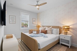 Un dormitorio con cama, mesitas de noche, ventilador de techo, televisor, ventana con persianas y decoración en la pared.