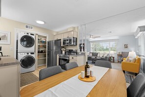 Eine moderne Küche mit integriertem Waschtrockner, einem Essbereich mit Holztisch sowie ein Wohnzimmer mit Sofa und gelbem Sessel.