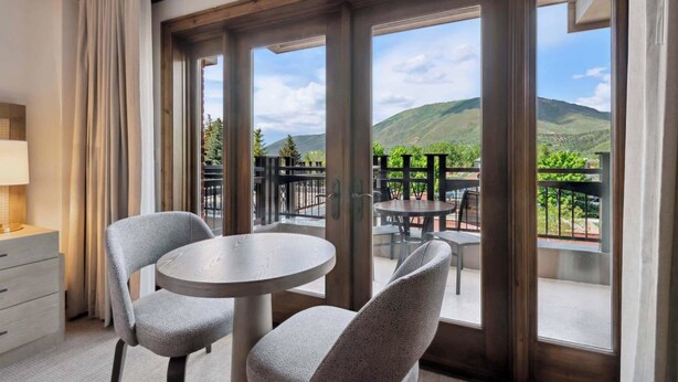 Une chambre d’hôtel avec un balcon, deux chaises, une table ronde et une vue sur des collines verdoyantes.