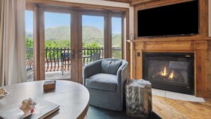 A room with a fireplace, a television, and a view of a balcony with outdoor furniture.