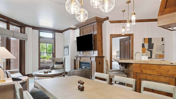 A modern kitchen with a large island, a built-in oven, and a flat-screen TV. There is a dining area with a long table and chairs, and a seating area with sofas and a coffee table.