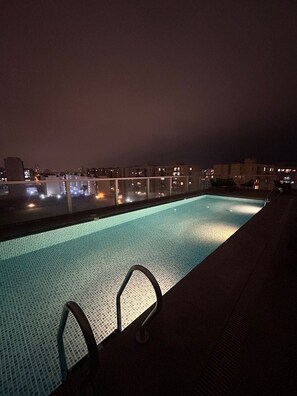 A rooftop swimming pool with a glass balustrade, surrounded by city buildings at night. - Beautiful Apartment in Lima (Lima)