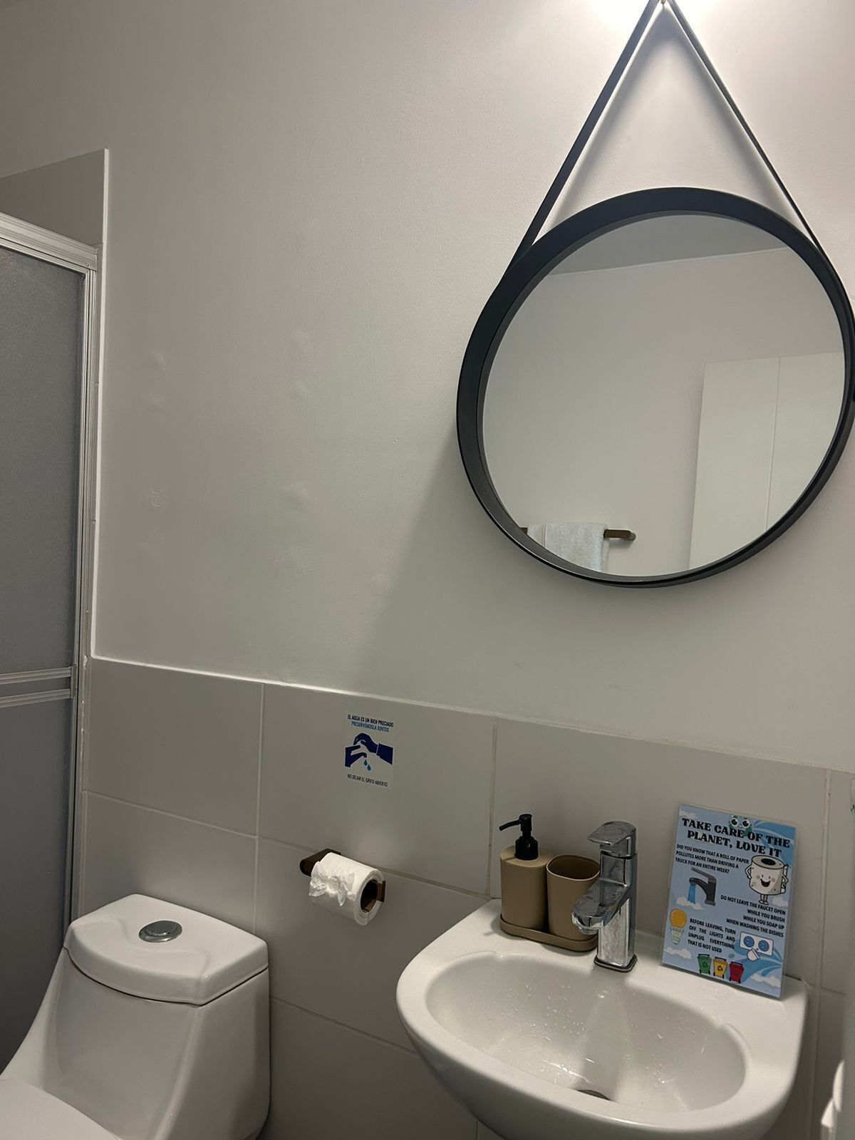 Ein Badezimmer mit Toilette, Waschbecken und einem Spiegel mit schwarzem Rahmen. An der Wand hängt ein Schild und neben der Toilette befindet sich eine Rolle Toilettenpapier.