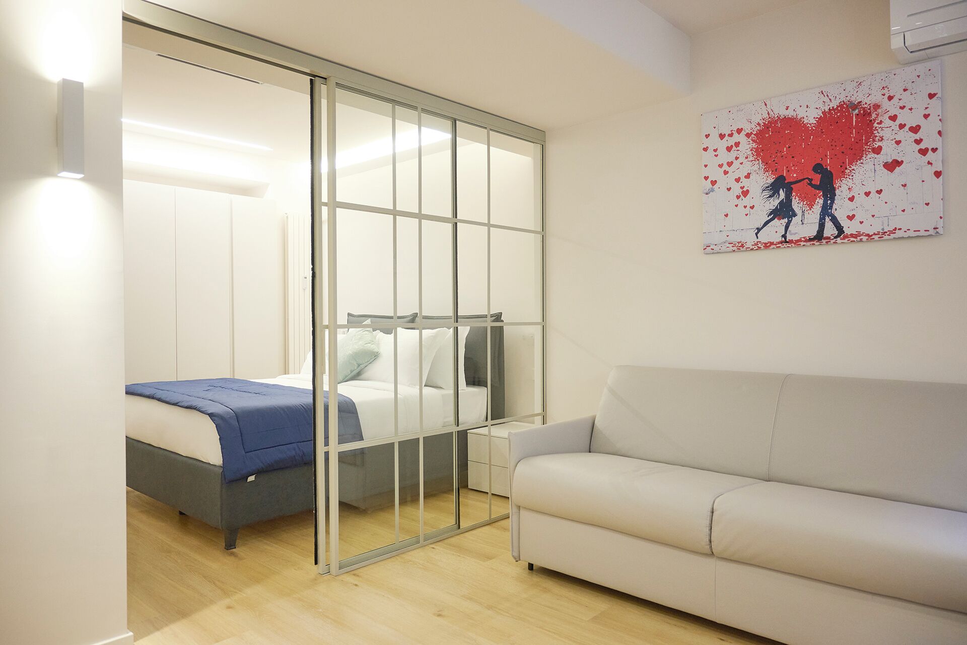 A bedroom with a bed, a sliding glass door, a white sofa, and a painting of two figures on a wall.