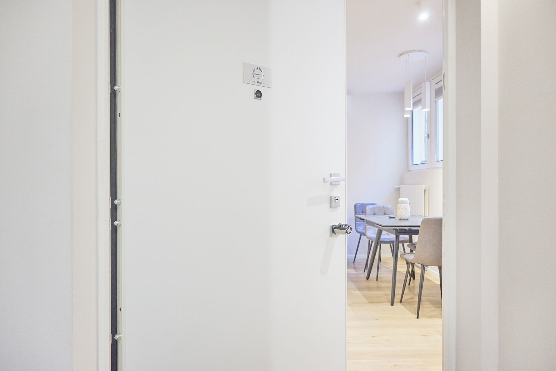 A modern hotel room with a white door, a small window, and a view of a dining area with a table and chairs.