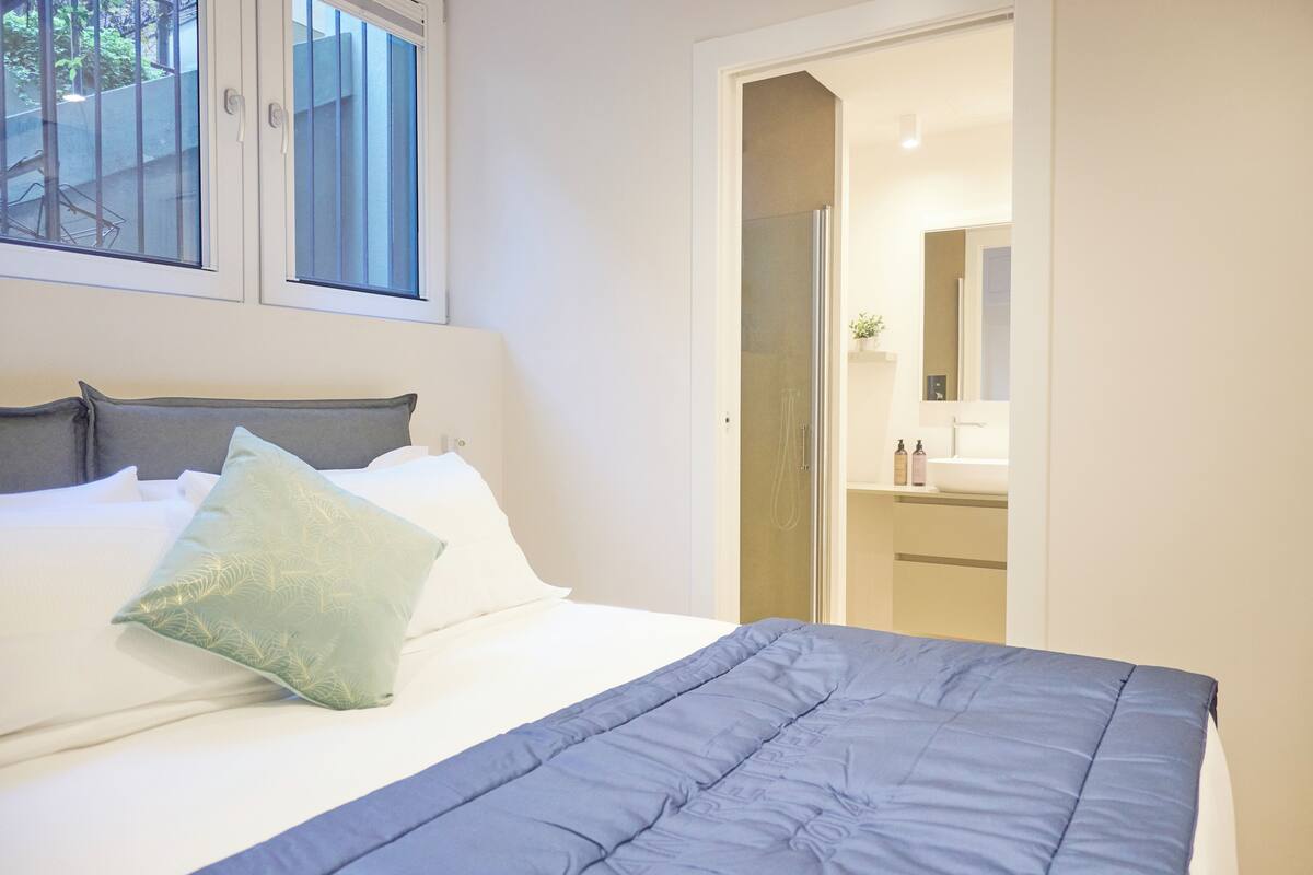 Un lit bien fait, avec une couverture bleue et un oreiller vert, une fenêtre avec des stores et une salle de bain visible à travers une porte ouverte.